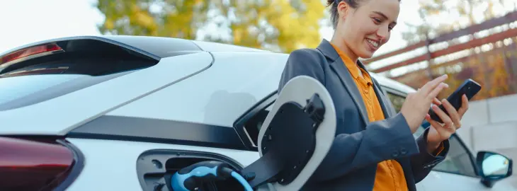 Woman charging an electric vehicle at home while checking her phone, illustrating smart energy use.