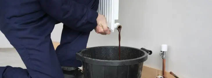 Heating engineer draining dark sludge from a radiator into a bucket during system cleaning.
